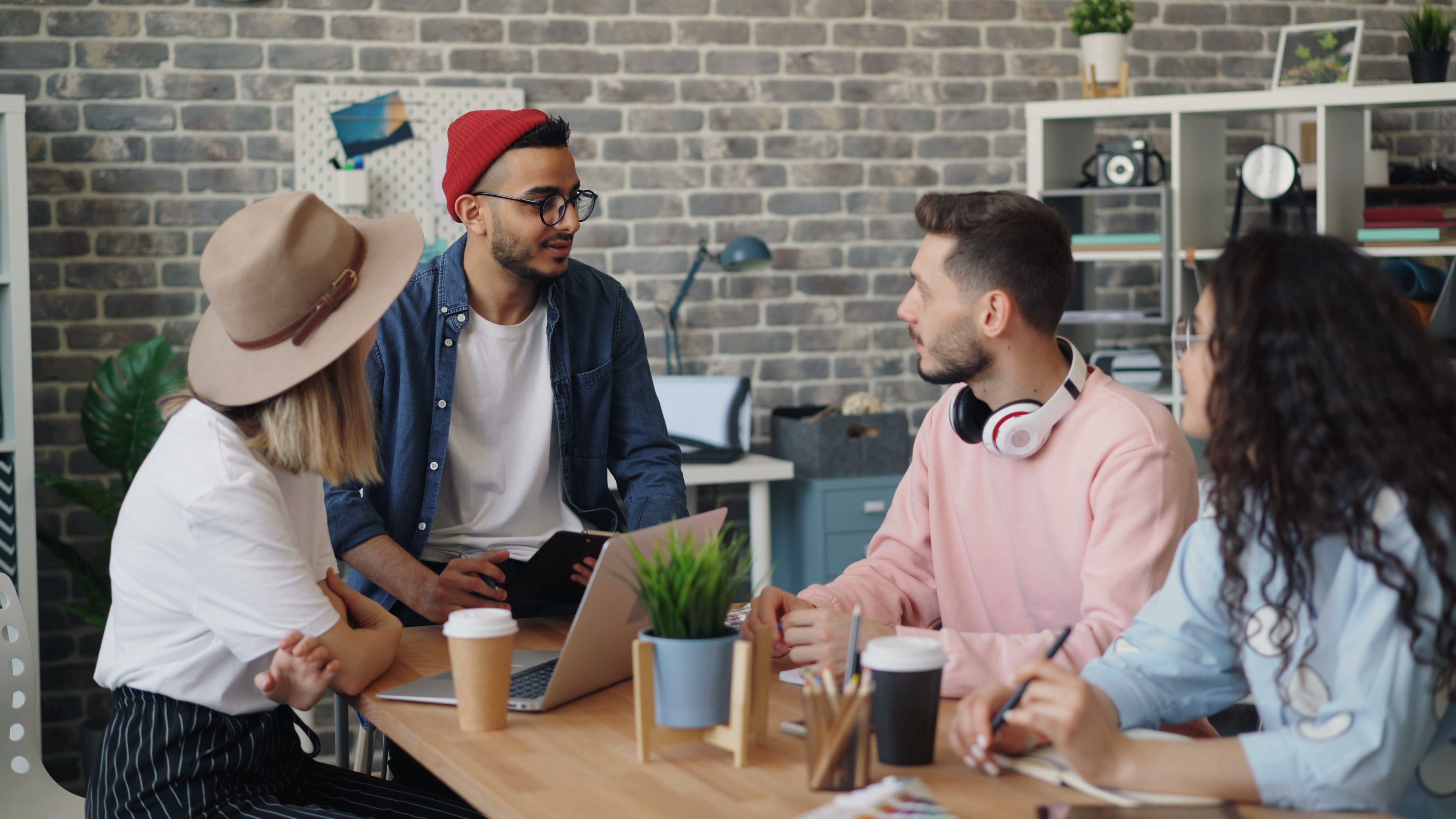 A group of four people sitting around a table in a casual meeting, with laptops, notebooks, and coffee cups in a modern office setting.