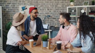A group of four people sitting around a table in a casual meeting, with laptops, notebooks, and coffee cups in a modern office setting.