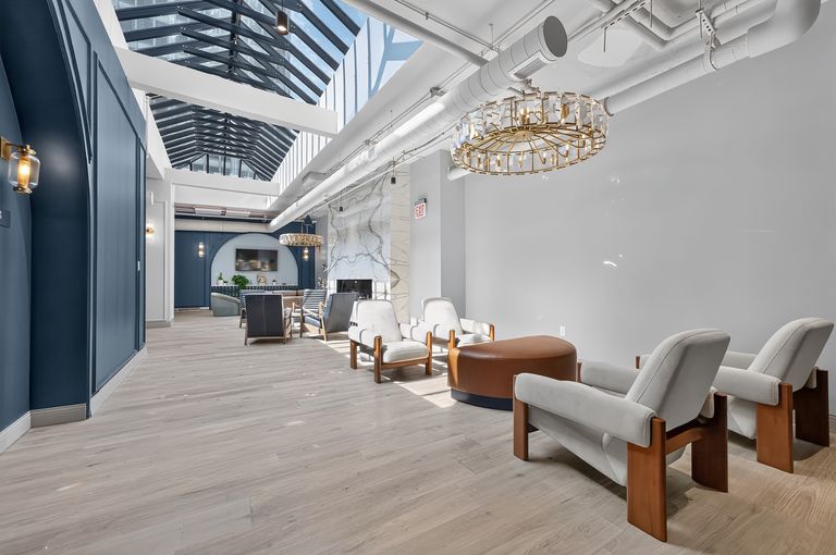 Modern lobby with skylight, gray chairs, round brown ottoman, and decorative chandeliers. Large windows cast natural light on wood flooring.
