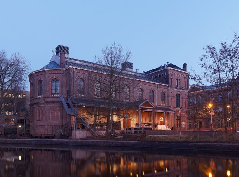 Historic brick building by a canal at dusk, with warm interior lights glowing through arched windows, reflecting in the water.