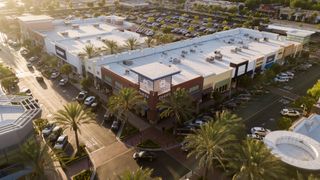 Aerial view of a shopping center with palm trees, multiple stores with flat roofs, and a parking lot filled with cars.
