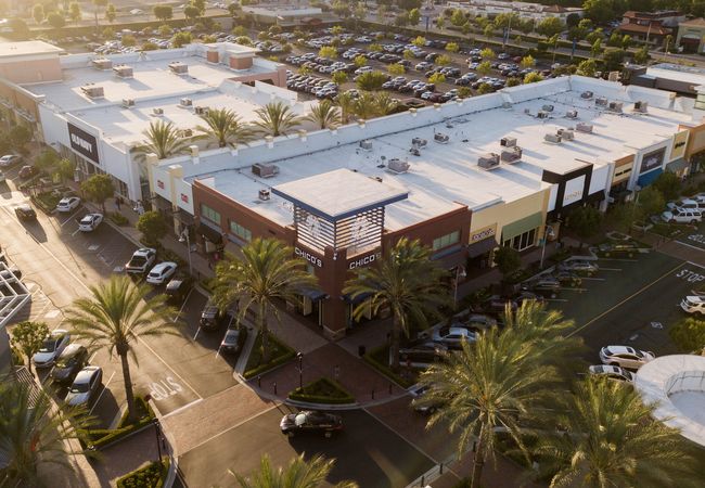 Aerial view of a shopping center with palm trees, multiple stores with flat roofs, and a parking lot filled with cars.