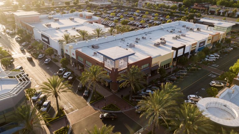 Aerial view of a shopping center with palm trees, multiple stores with flat roofs, and a parking lot filled with cars.