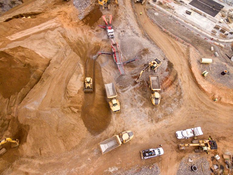 Aerial view of a construction site with several yellow excavators, dump trucks, and a white truck on dirt ground, surrounded by earth and equipment.