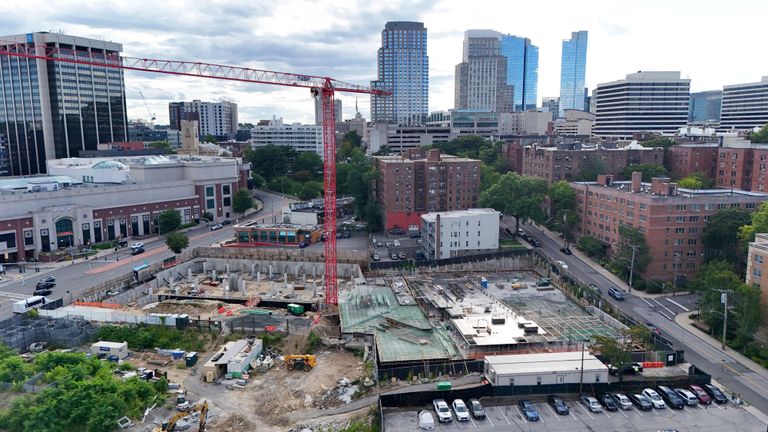 Aerial view of a construction site with a red crane amidst urban buildings and skyscrapers under a cloudy sky.