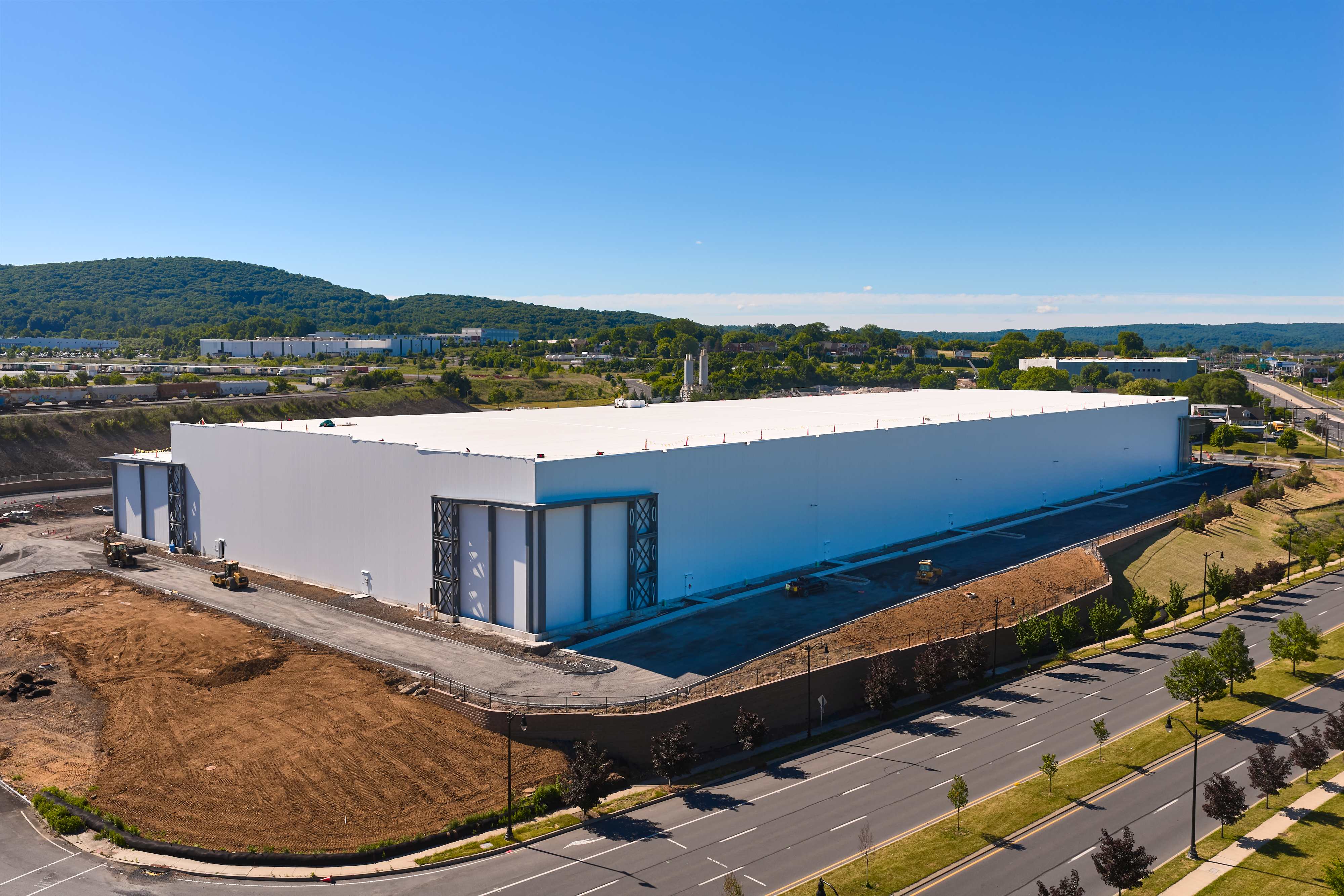 Large white warehouse building under blue sky, surrounded by roads and green landscape, with hills in the background.