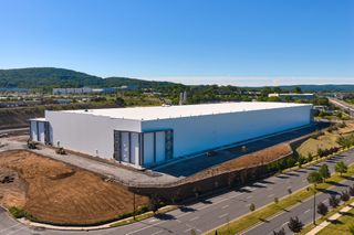 Large white warehouse building under blue sky, surrounded by roads and green landscape, with hills in the background.