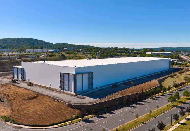Large white warehouse building under blue sky, surrounded by roads and green landscape, with hills in the background.