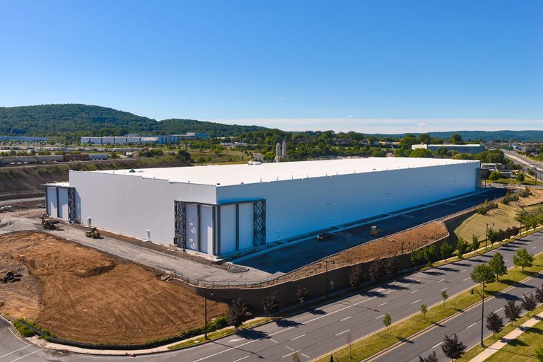 Large white warehouse building under blue sky, surrounded by roads and green landscape, with hills in the background.