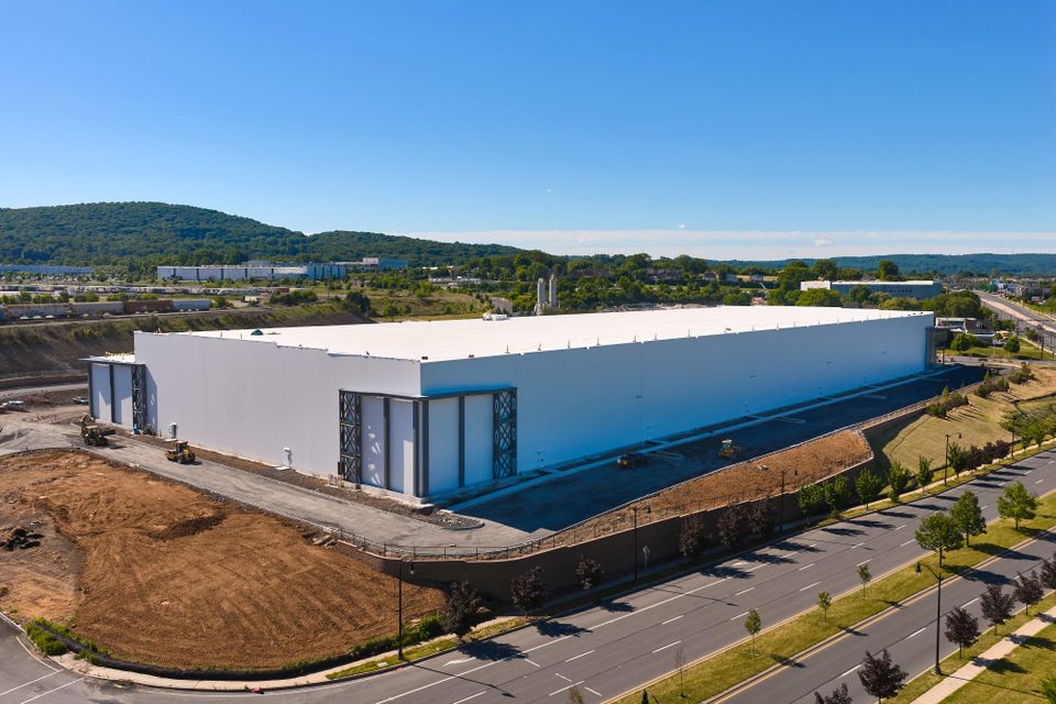 Large white warehouse building under blue sky, surrounded by roads and green landscape, with hills in the background.