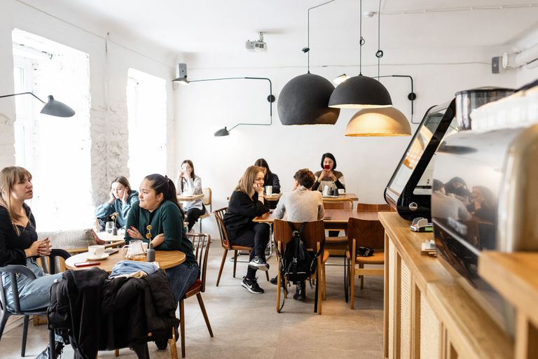 People sitting and talking at tables inside a cozy, modern cafe with large pendant lights and natural light coming through the windows.