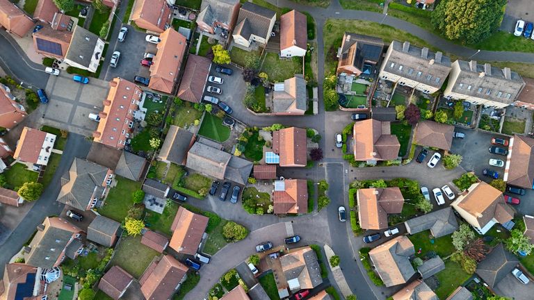 Aerial view of a suburban neighborhood with houses, driveways, parked cars, green lawns, and trees lining winding streets.