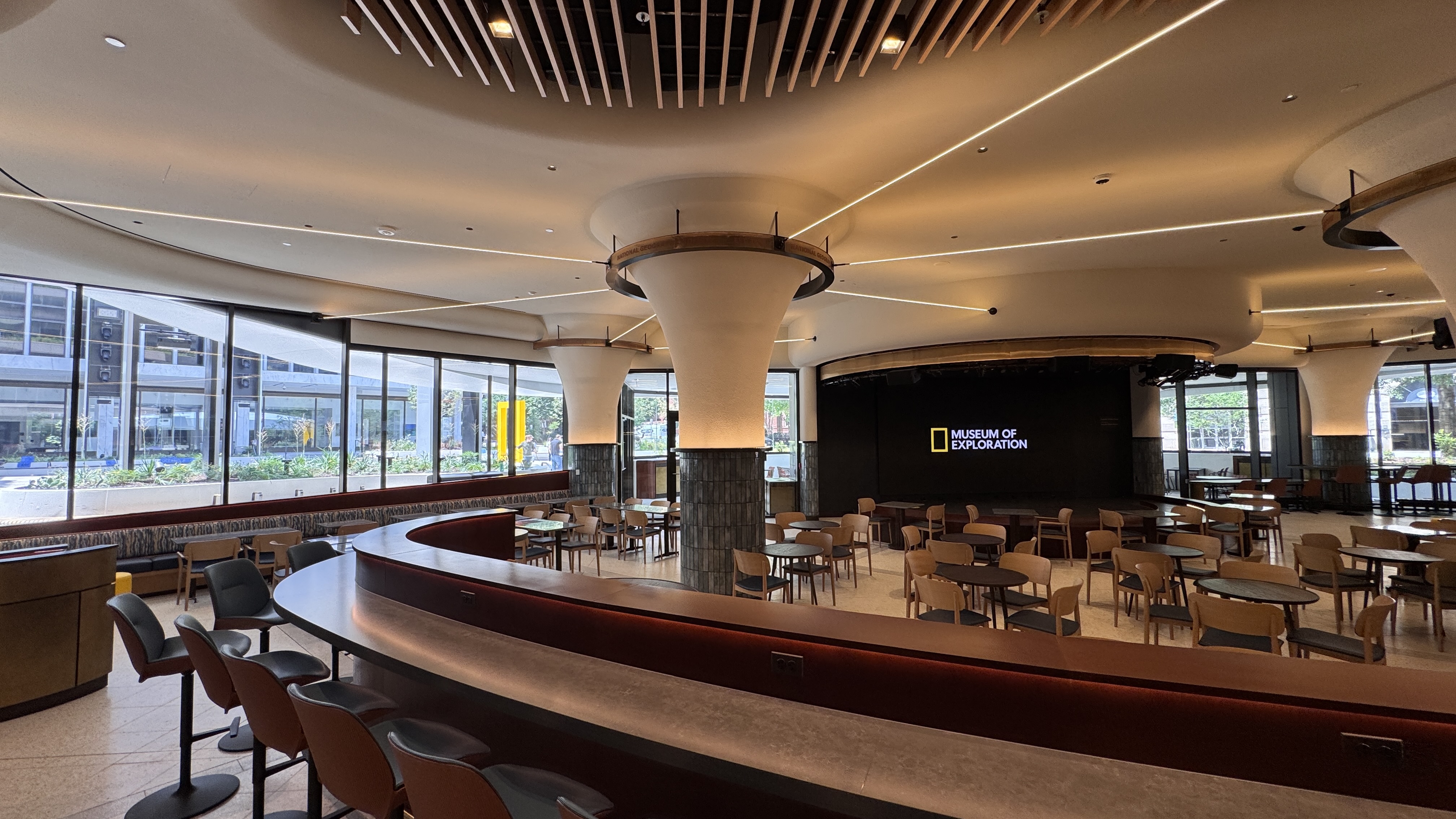 Modern museum cafe with round tables, cushioned chairs, and large windows. Wall displays "Museum of Exploration". Minimalist ceiling design.