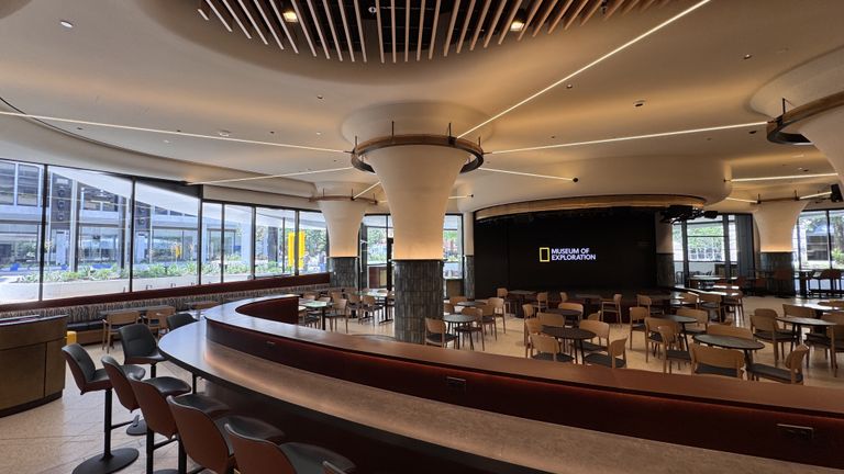 Modern museum cafe with round tables, cushioned chairs, and large windows. Wall displays "Museum of Exploration". Minimalist ceiling design.