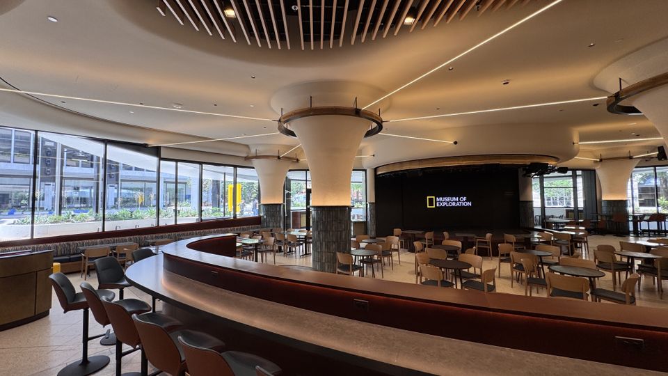 Modern museum cafe with round tables, cushioned chairs, and large windows. Wall displays "Museum of Exploration". Minimalist ceiling design.