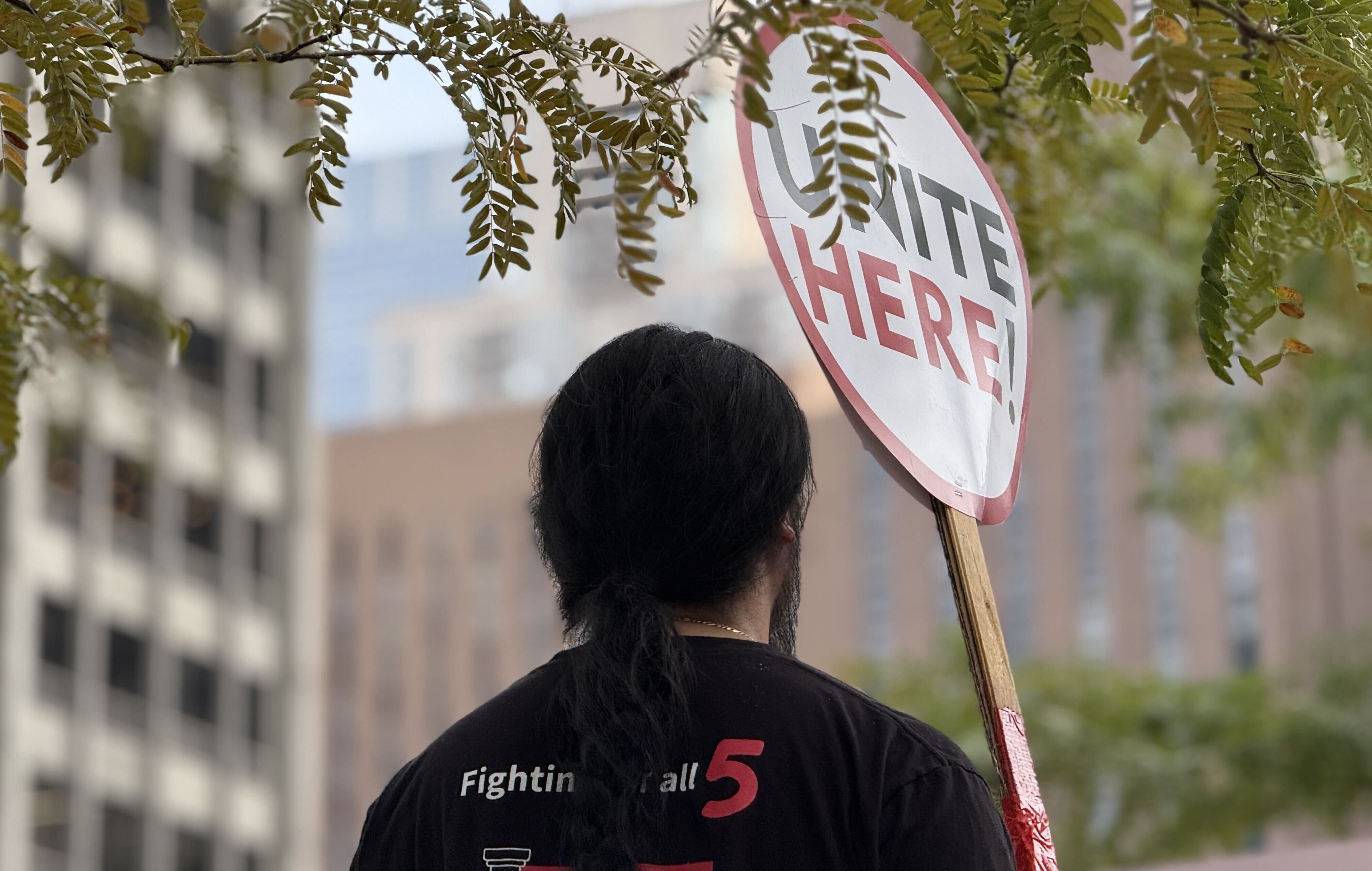 A person holding a sign reading "UNITE HERE!" stands under tree branches, with blurred buildings in the background.