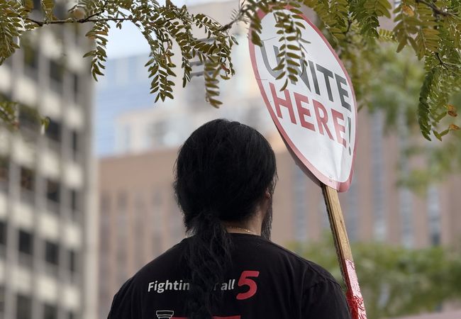 A person holding a sign reading "UNITE HERE!" stands under tree branches, with blurred buildings in the background.