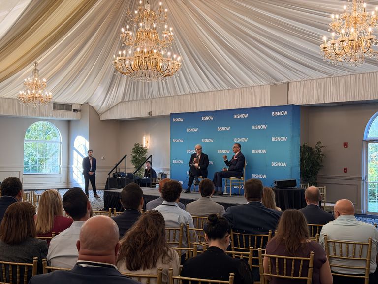 Audience seated while two men speak on stage at a Bisnow event in an elegant room with chandeliers and arched windows.