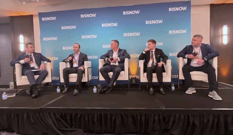 Five men in business attire sit on stage chairs, engaged in a panel discussion at a Bisnow event, with a branded backdrop behind them.