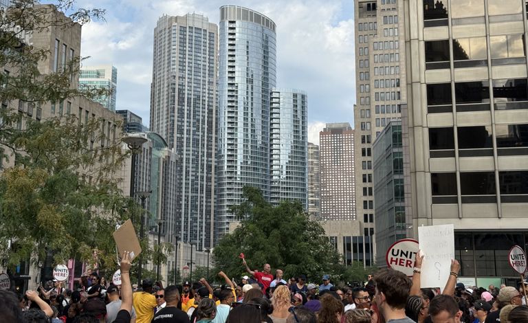 Crowd gathered in a city street protest, holding signs amid tall buildings under a partly cloudy sky.
