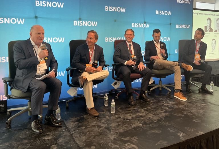 Five men in business attire sit on a panel discussion stage with a BISNOW backdrop and a presentation screen displaying speaker names.