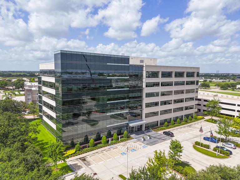 Modern office building with reflective glass windows, surrounded by landscaped greenery and a parking area under a partly cloudy blue sky.