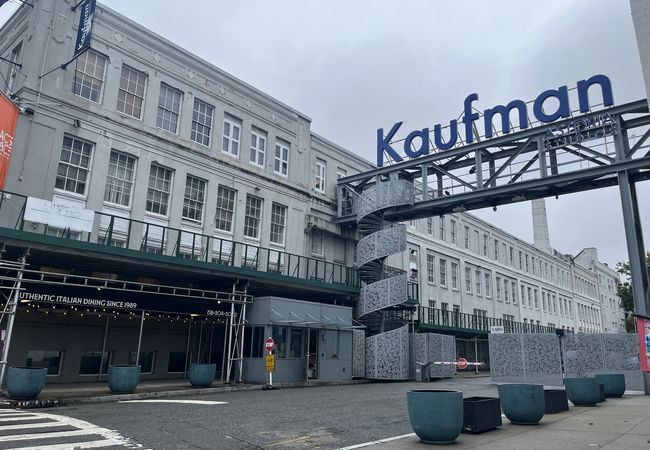 Exterior of a large, industrial-style building with a sign reading "Kaufman Astoria Studios" and a modern spiral staircase. Overcast sky.