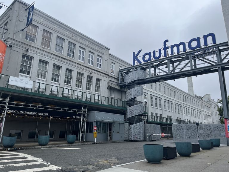 Exterior of a large, industrial-style building with a sign reading "Kaufman Astoria Studios" and a modern spiral staircase. Overcast sky.