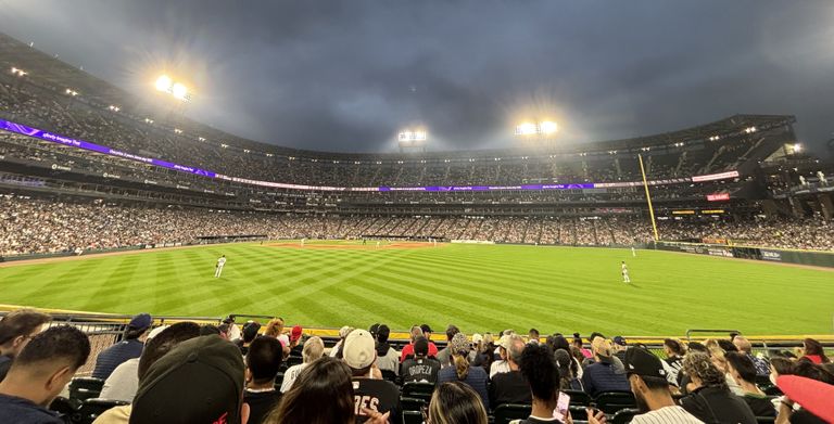 A crowded baseball stadium at night with bright lights illuminating the green field and overcast sky, viewed from the outfield stands.