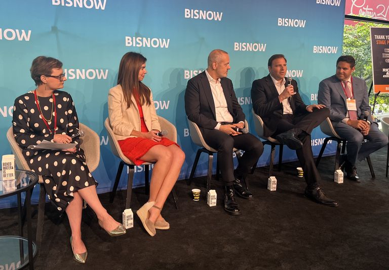 Five people seated on a stage speak into microphones at a Bisnow event. A blue backdrop with the Bisnow logo is behind them.
