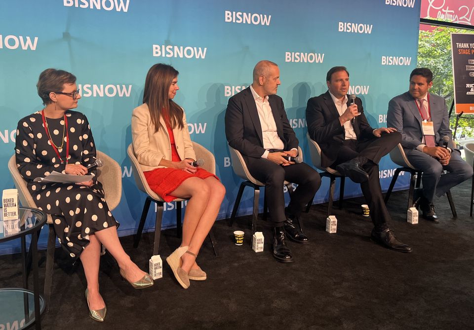 Five people seated on a stage speak into microphones at a Bisnow event. A blue backdrop with the Bisnow logo is behind them.