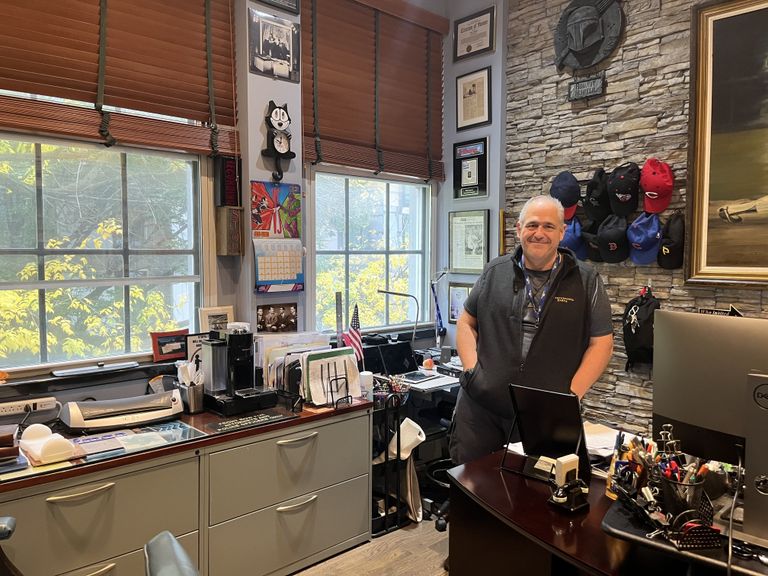 A person smiling while standing in a cluttered office with various decor, two large windows, and a collection of baseball caps on a stone wall.