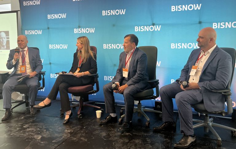 Four people in business attire are seated on a panel at a BISNOW event, engaging in a discussion while holding microphones and notes.