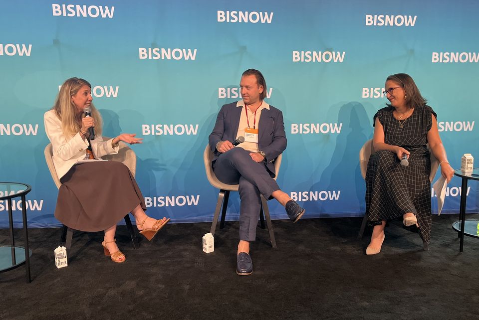 Three people seated on a stage with a "Bisnow" backdrop, engaged in a panel discussion. One person holds a microphone.