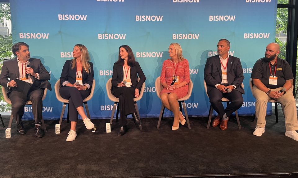 Six panelists sitting on stage with "BISNOW" backdrop, engaged in discussion, wearing business attire with lanyards.