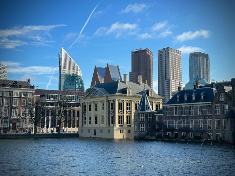 Historic and modern buildings by a canal in The Hague under a bright blue sky with scattered clouds, showcasing diverse architecture.