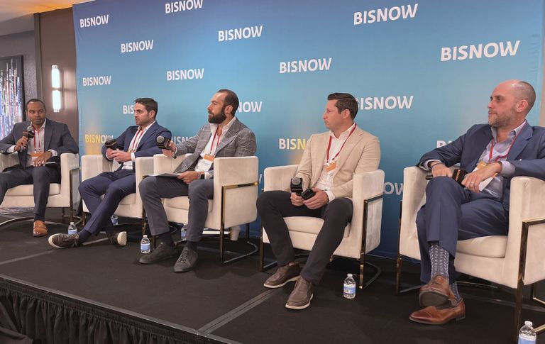 Five men in suits sit on a panel stage at a Bisnow event, holding microphones and engaging in discussion.