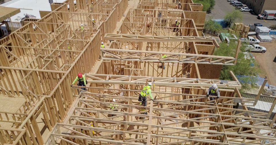 Construction workers in safety gear are assembling a wooden framework for a building. Numerous beams and partially built walls are visible.