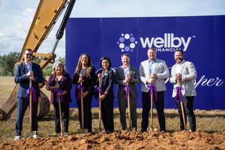 A group of seven people, holding shovels with purple ribbons, participate in a groundbreaking ceremony in front of Wellby Financial signage.