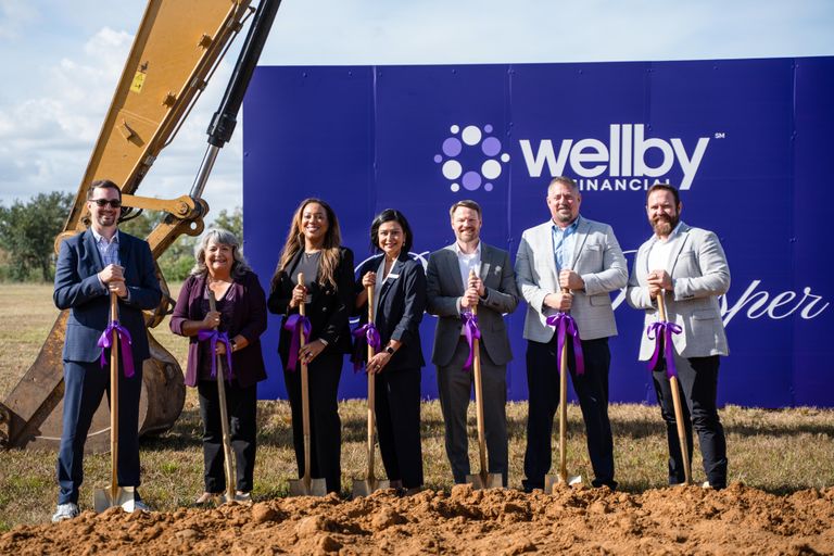 A group of seven people, holding shovels with purple ribbons, participate in a groundbreaking ceremony in front of Wellby Financial signage.