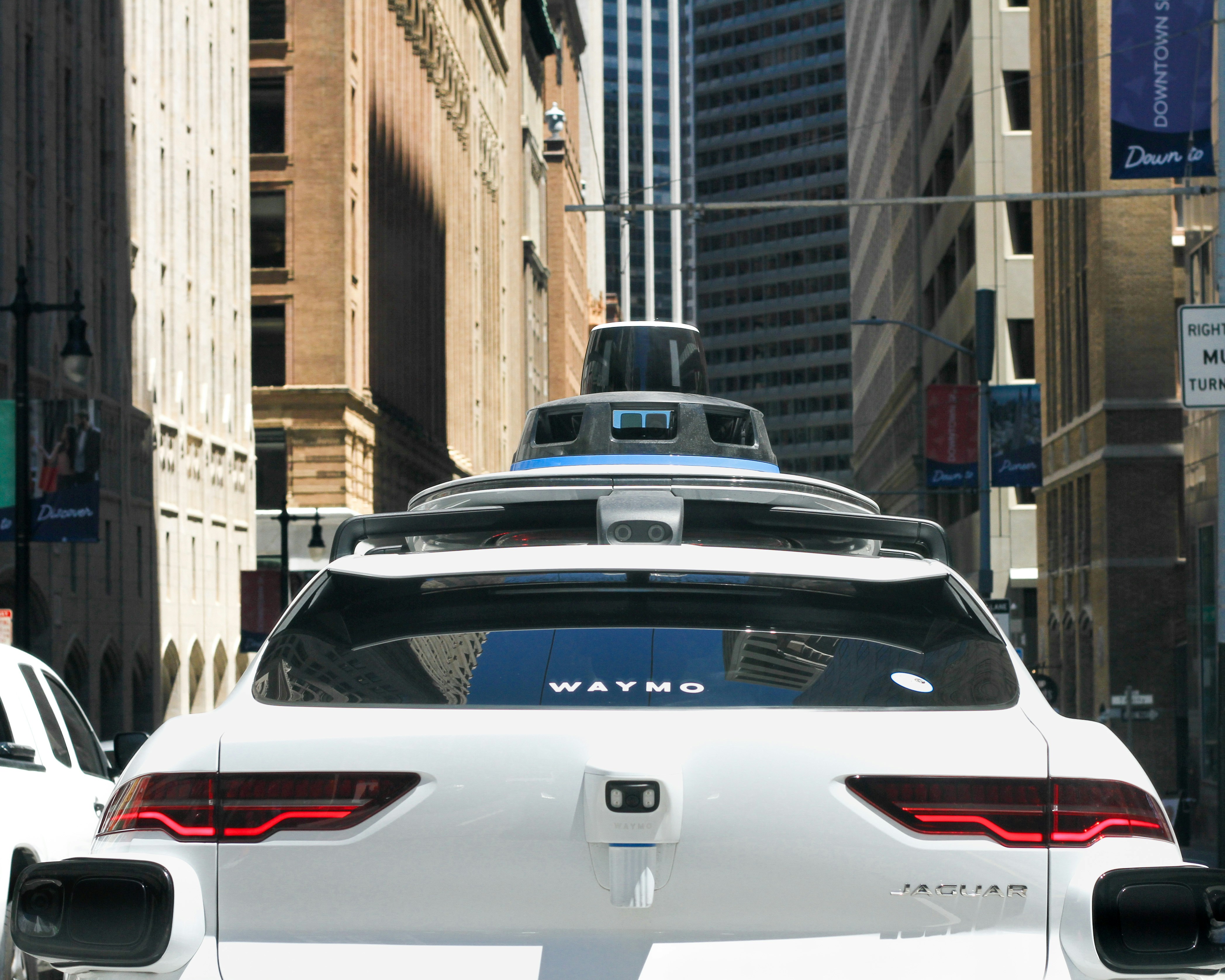 A white Waymo self-driving car navigates a city street, surrounded by tall buildings and traffic, with sensors mounted on the roof.