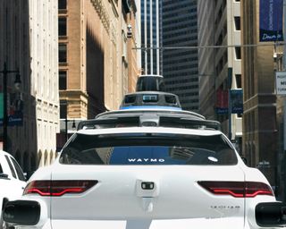 A white Waymo self-driving car navigates a city street, surrounded by tall buildings and traffic, with sensors mounted on the roof.