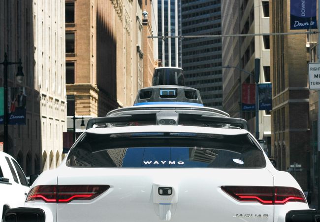 A white Waymo self-driving car navigates a city street, surrounded by tall buildings and traffic, with sensors mounted on the roof.