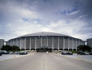 A large domed stadium with a concrete exterior stands under a cloudy sky, surrounded by a parking lot with a few parked cars.