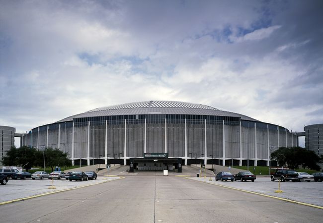 A large domed stadium with a concrete exterior stands under a cloudy sky, surrounded by a parking lot with a few parked cars.