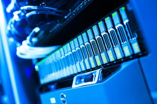 Close-up of a row of server racks with multiple hard drives, illuminated by blue light, and network cables in the background.