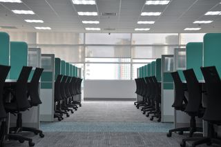 Empty office space with rows of black swivel chairs and turquoise cubicle dividers, under bright fluorescent lighting.