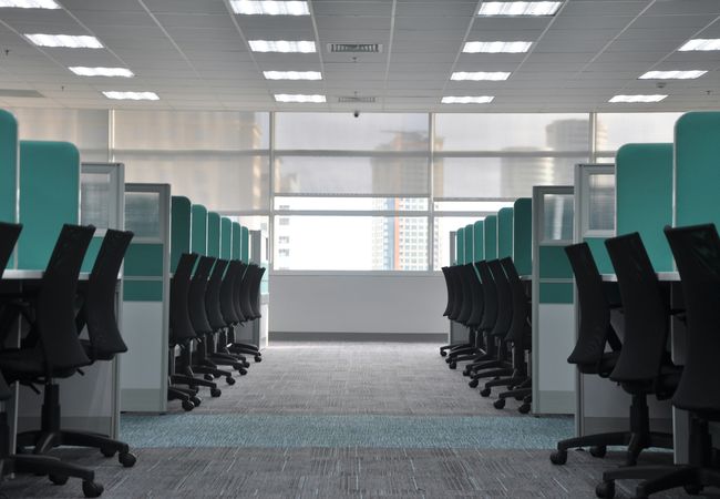 Empty office space with rows of black swivel chairs and turquoise cubicle dividers, under bright fluorescent lighting.