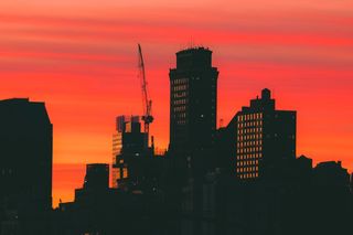 Silhouette of city buildings and a crane against a vivid orange and red sunset sky.