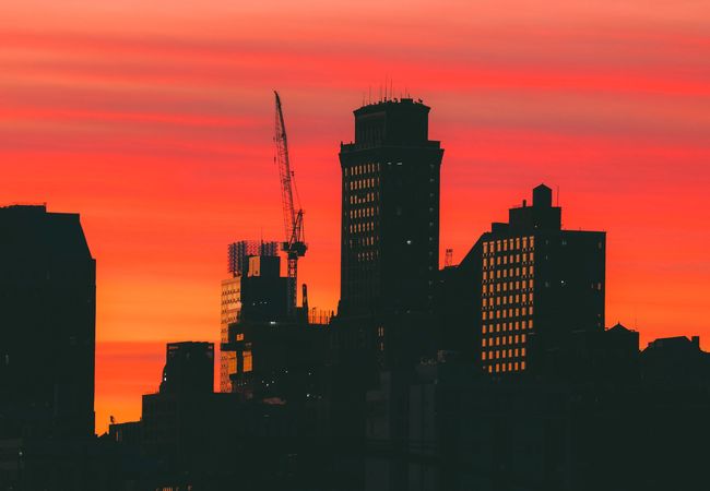 Silhouette of city buildings and a crane against a vivid orange and red sunset sky.